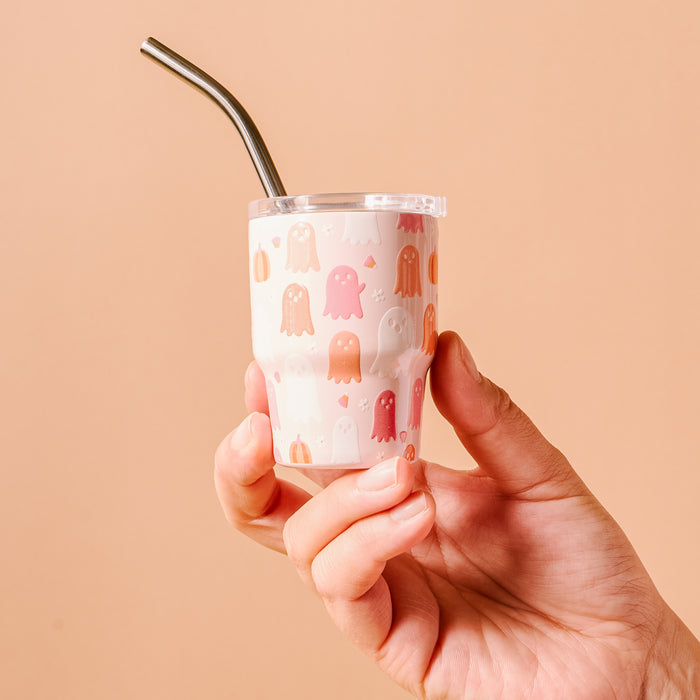 A hand holds The Darling Effect's Ghost Party 2 oz Tiny Tumbler, featuring a cute pastel ghost design on a light background with a metal straw, set against a peach-colored backdrop.