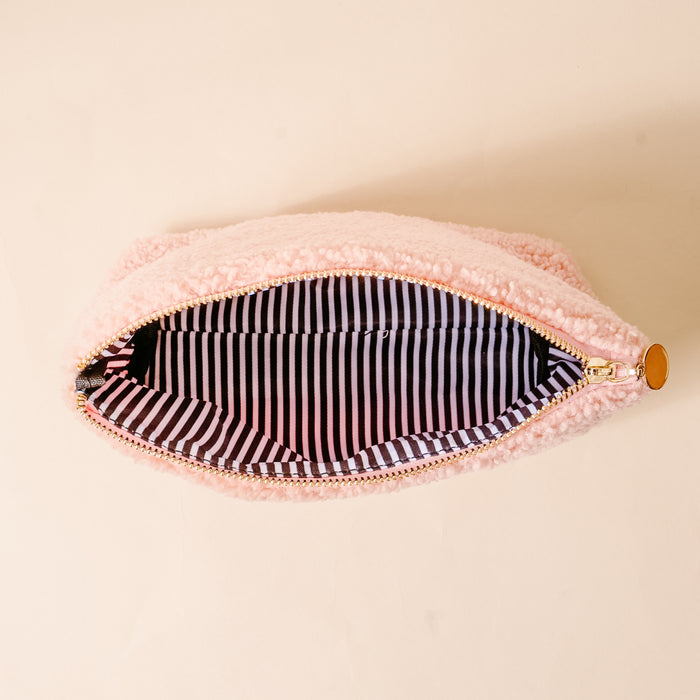 The Darling Effect’s Ghosts & Pearls Teddy Pouch, crafted from cozy pink sherpa with a gold zipper pull, is shown open to reveal its black and white striped lining—a charming fall accessory resting on a beige surface.