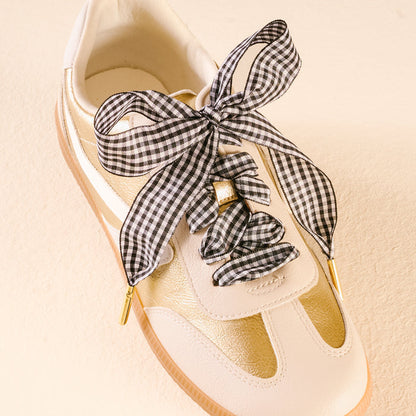 The Darling Effect’s Gingham Black Step It Up Shoelaces, tied in a large bow, are shown on a cream and gold sneaker against a light beige background.