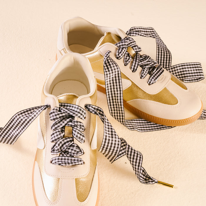A pair of white and gold sneakers with The Darling Effect's Gingham Black Step It Up Shoelaces, shown on a cream-colored surface.