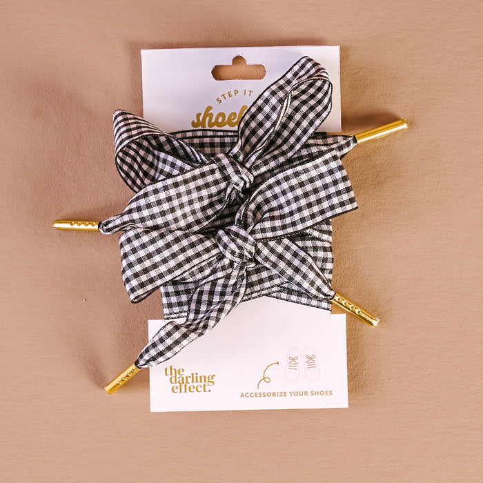 Gingham Black Step It Up Shoelaces by The Darling Effect feature black and white gingham bows on gold shoe clips, ideal for accessorizing sneakers. Displayed on white packaging with "the darling effect" text against a beige background.