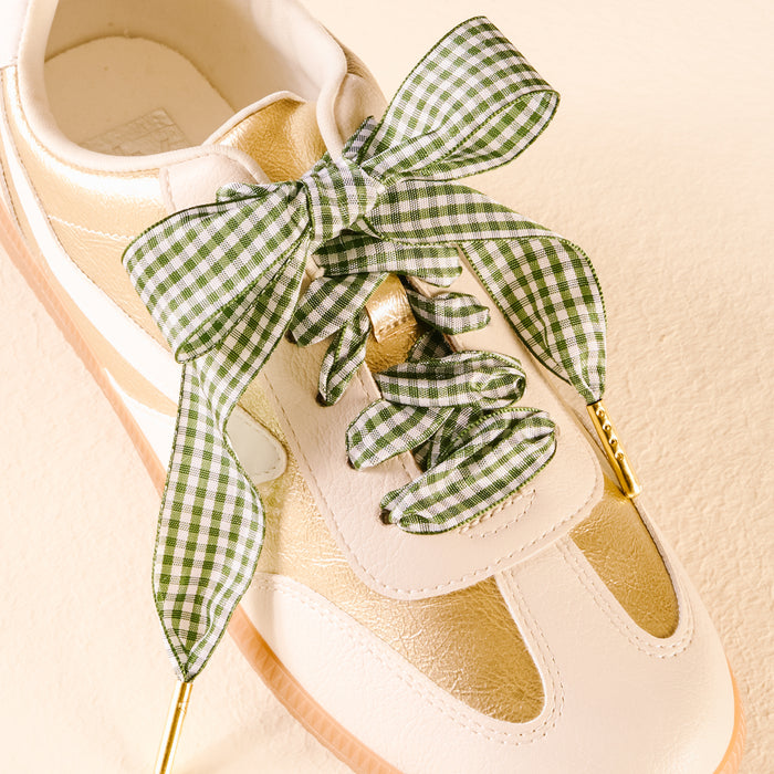 A white and gold sneaker with The Darling Effect's Gingham Green Step It Up Shoelaces, featuring durable green and white gingham tied in a large bow, is set against a light beige background.