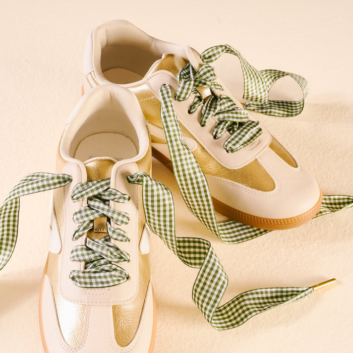 A pair of cream and gold sneakers with The Darling Effect's Gingham Green Step It Up Shoelaces, featuring a green and white gingham pattern, displayed on a light beige surface.