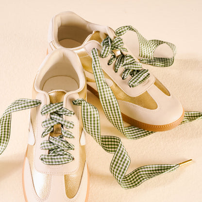 A pair of cream and gold sneakers with The Darling Effect's Gingham Green Step It Up Shoelaces, featuring a green and white gingham pattern, displayed on a light beige surface.