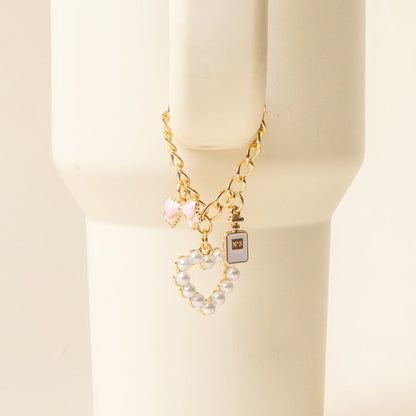 The Glamour Tumbler Charm by the-darling-effect features a gold chain bracelet with three charms—a pink bow, black perfume bottle, and pearl heart—on a cream stand, perfect for pairing with your favorite tumbler accessories.