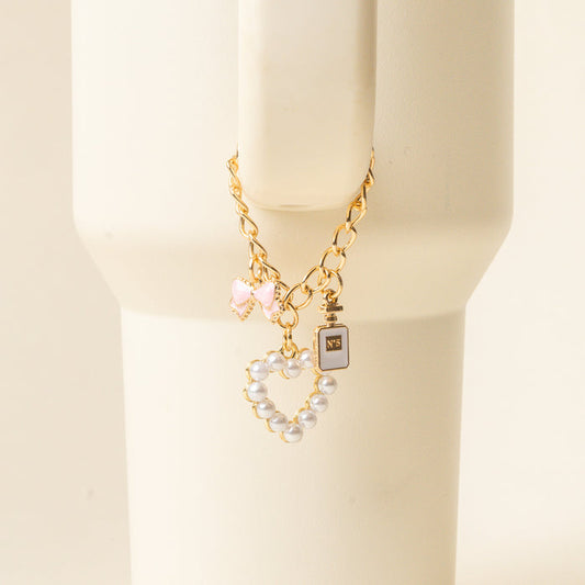 The Glamour Tumbler Charm by the-darling-effect features a gold chain bracelet with three charms—a pink bow, black perfume bottle, and pearl heart—on a cream stand, perfect for pairing with your favorite tumbler accessories.