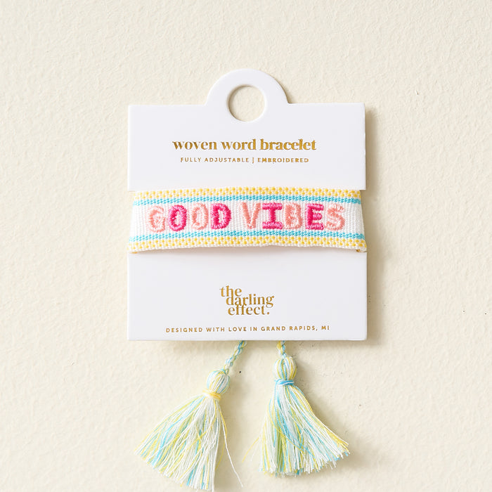 The Darling Effect’s Good Vibes Woven Bracelet features pastel blue, yellow, and white stripes with “GOOD VIBES” in pink embroidery, adjustable fit, tassels at the bottom, and comes displayed on a white card.