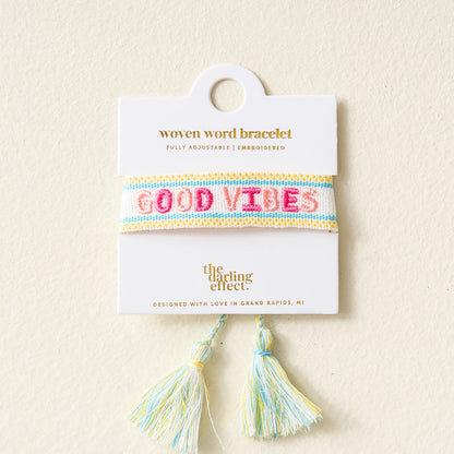 The Darling Effect’s Good Vibes Woven Bracelet features pastel blue, yellow, and white stripes with “GOOD VIBES” in pink embroidery, adjustable fit, tassels at the bottom, and comes displayed on a white card.