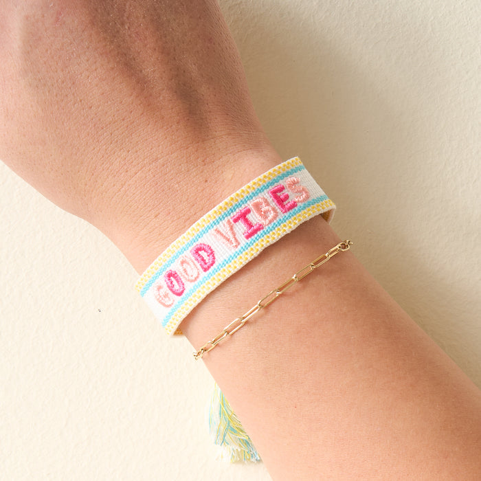 The Darling Effect's Good Vibes Woven Bracelet features white adjustable fabric with "GOOD VIBES" embroidered in pink and light blue—perfect for stacking with a gold chain for a trendy look.