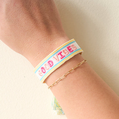 The Darling Effect's Good Vibes Woven Bracelet features white adjustable fabric with "GOOD VIBES" embroidered in pink and light blue—perfect for stacking with a gold chain for a trendy look.