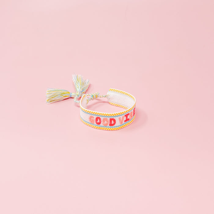 The Darling Effect's Good Vibes Woven Bracelet features pastel embroidery and playful tassels, with "GOOD VIBES" in pink and orange lettering.