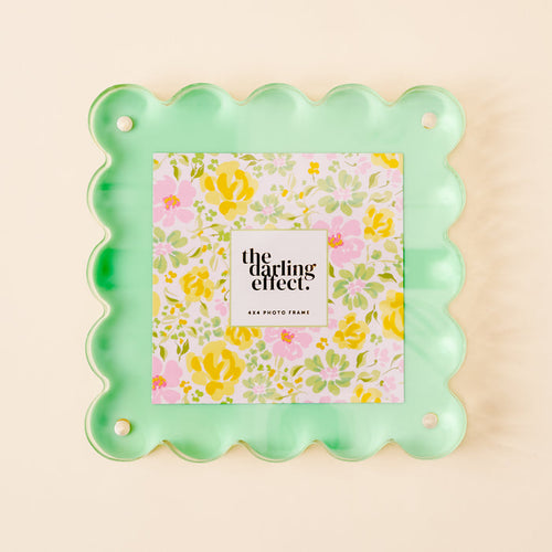 The Green Square Scalloped Acrylic Frame by the-darling-effect features mint green scalloped edges and displays a floral print with pink, yellow, and green accents. The center says "the darling effect. 4x4 photo frame" on a white square over pale beige.