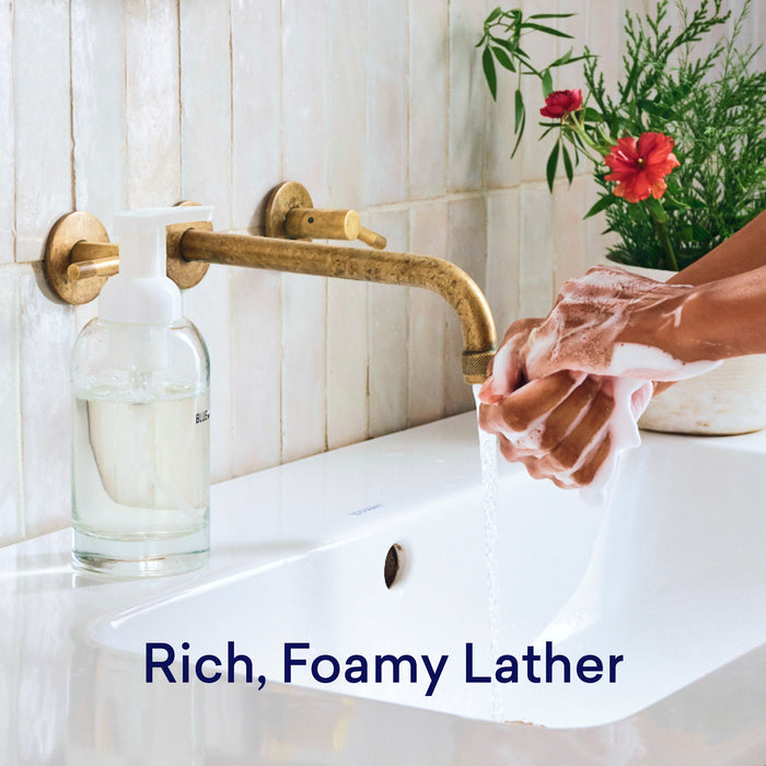 A person washes their hands under a brass faucet, creating a rich, foamy lather with the Hand Soap Starter 2-Pack’s plastic-free tablets, beside flowers in a vase on a white bathroom sink. Text: Rich, Foamy Lather.