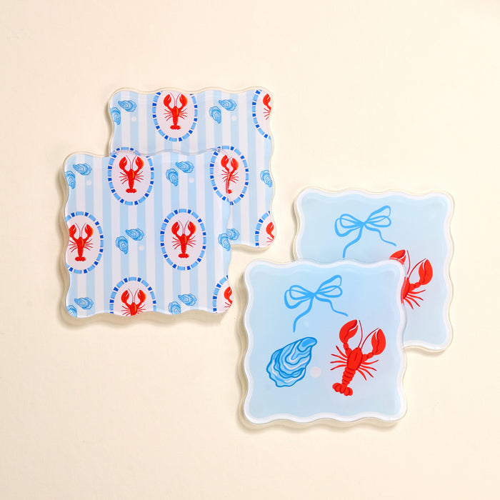 The Darling Effect’s Harbor House Table Talk Acrylic Coaster Set features four scalloped-edge coasters: two with red lobsters and blue shells on stripes, and two with a blue oyster, bow, and lobsters on a light blue background.