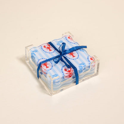 The Darling Effect’s Harbor House Table Talk Acrylic Coaster Set features scallop-edged coasters with blue stripes and red lobster prints in a clear holder, tied with a navy ribbon—perfect table decor for any occasion.