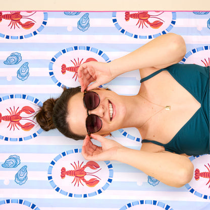 A smiling woman in sunglasses and a teal swimsuit relaxes on the Harbor House XL Quick-Dry Beach Towel by The Darling Effect, featuring red lobsters and blue seashells, looking up at the camera.