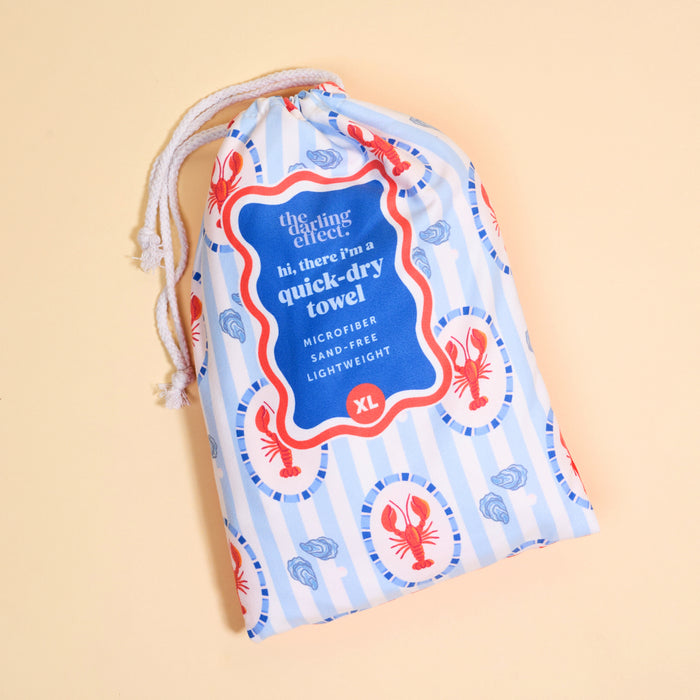 The Darling Effect Harbor House XL Quick-Dry Beach Towel comes in a blue and white striped drawstring bag with red lobsters. It's microfiber, sand-free, lightweight—perfect for all your adventures.