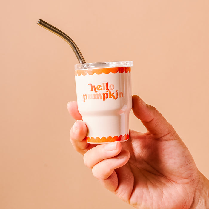 A hand holds The Darling Effect's Hello Pumpkin 2 oz Tiny Tumbler, a white cup with orange scalloped edges and "hello pumpkin" lettering. This mini tumbler is a charming autumn party favor set against a plain beige background.
