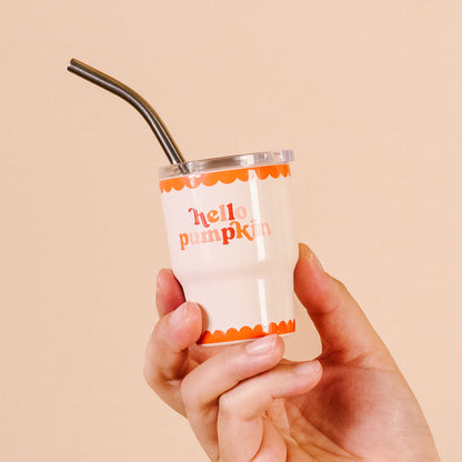 A hand holds The Darling Effect’s Hello Pumpkin 2 oz Tiny Tumbler, featuring white with orange scalloped trim and “hello pumpkin” in pink and orange lettering, plus a metal straw—ideal for autumn party favors.