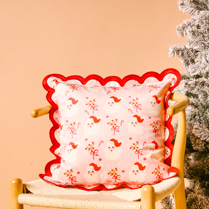 The Darling Effect’s Holly Jolly Santa Scalloped Square Throw Pillow adds festive flair to a wooden chair next to a frosted Christmas tree, featuring red scalloped edges and playful Santa, bow, and holly patterns.