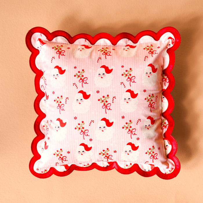 The Darling Effect's Holly Jolly Santa Scalloped Square Throw Pillow features red scalloped edges and white fabric with Santa faces, candy canes, holly, and red bows for a cozy festive touch on any peach-colored background.