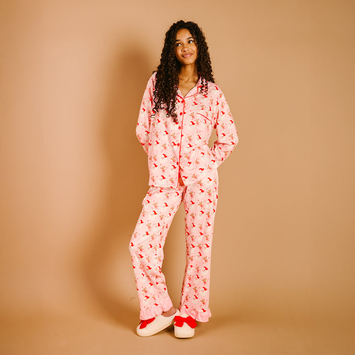 A woman with long curly hair stands against a tan background, wearing The Darling Effect’s Holly Jolly Santa Sweet Dreams Pajama Pants Set in light pink with red florals and matching slippers with red bows, smiling off to the side.