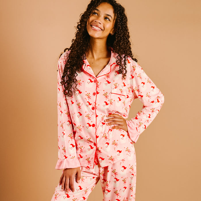 A woman with long curly hair smiles upward while wearing The Darling Effect's Holly Jolly Santa Sweet Dreams Pajama Pants Set in light pink with a red floral pattern, standing against a plain beige background.