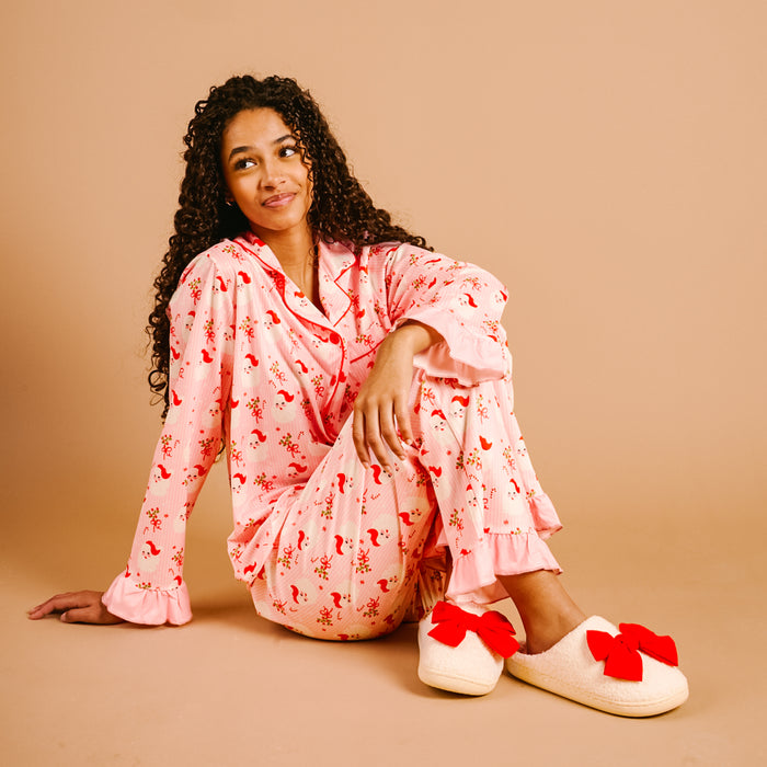 A woman with long curly hair sits on the floor in The Darling Effect's Holly Jolly Santa Sweet Dreams Pajama Pants Set—pink with a red floral pattern and ruffled cuffs—plus cream slippers with red bows, all in ultra-soft fabric against a beige background.