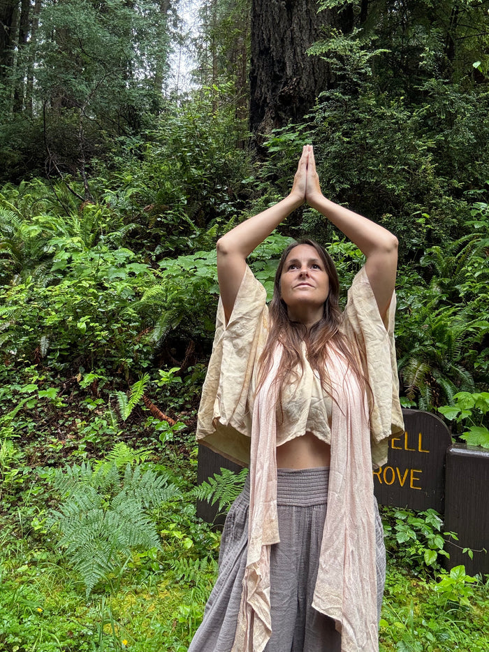 A woman in a forest setting with her hands pressed together above her head, wearing a beige open-front cardigan over a matching beige crop top and a gray skirt.