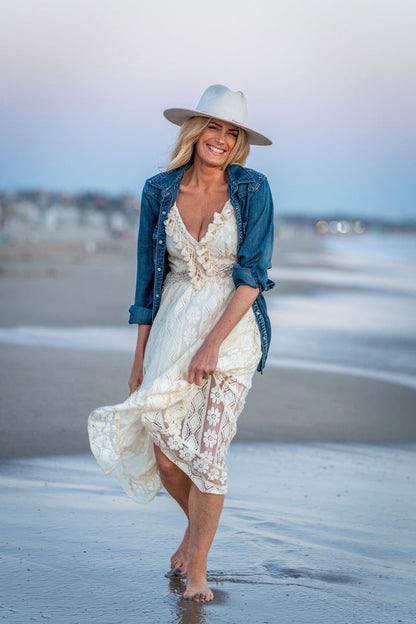 A woman walking barefoot on a beach wearing a white lace dress under a blue denim jacket and a wide-brimmed white hat.