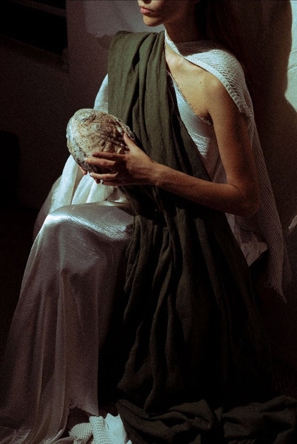 A model wearing a draped white dress with a dark olive green shawl, holding a textured shell in a dimly lit, moody setting.