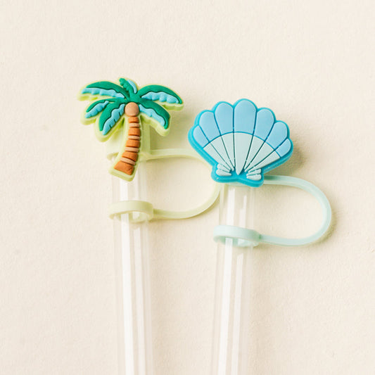 The Island Escape Straw Topper Set by the-darling-effect includes two clear reusable straws with leak-resistant silicone toppers—a green palm tree and a blue seashell—each attached with matching loops, ideal for brightening up any drink.