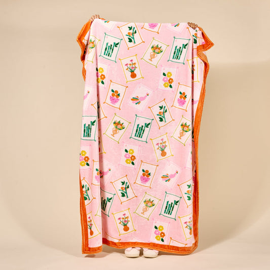 A person holds up The Darling Effect's It's Mahj Time Snuggle Up Plush Blanket, featuring vibrant flower vase designs and an orange border. Only their hands and white shoes show behind the cozy throw.