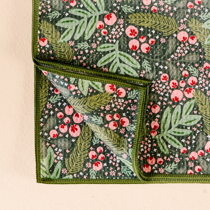 A close-up of The Darling Effect's Jolly Sprig Waffle Tea Towel shows a folded corner, highlighting its colorful botanical pattern with green leaves, red berries, and pink flowers on both sides.