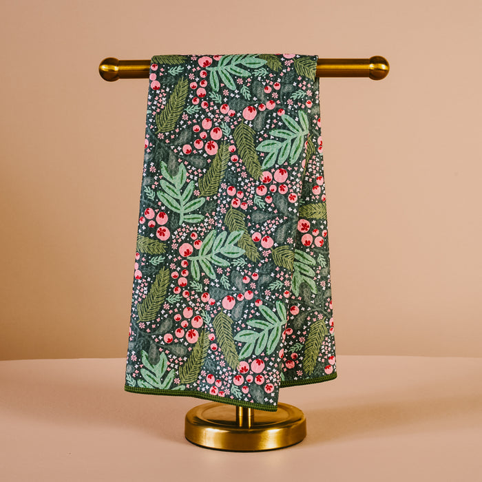The Darling Effect’s Jolly Sprig Waffle Tea Towel, featuring a dark green waffle weave with colorful leaves and red berries, is displayed on a gold towel stand against a beige background.