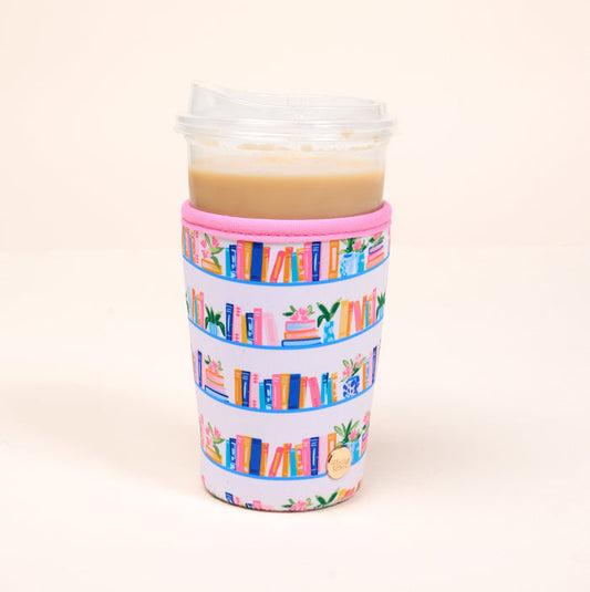 The Darling Effect’s Large-Book Shelfie Sip Savvy Drink Sleeve, featuring colorful bookshelf and plant designs, wraps around a clear cup with a light brown iced drink and lidded top, set against a plain light background.
