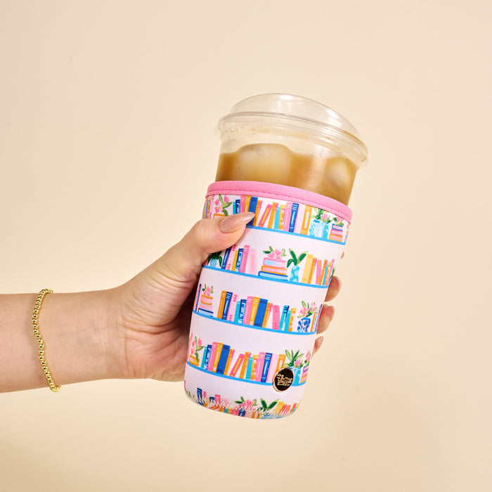 A hand holds an iced coffee in a clear plastic cup with The Darling Effect’s Large-Book Shelfie Sip Savvy Drink Sleeve, featuring illustrated books and plants on a cream background. The person wears a gold chain bracelet.