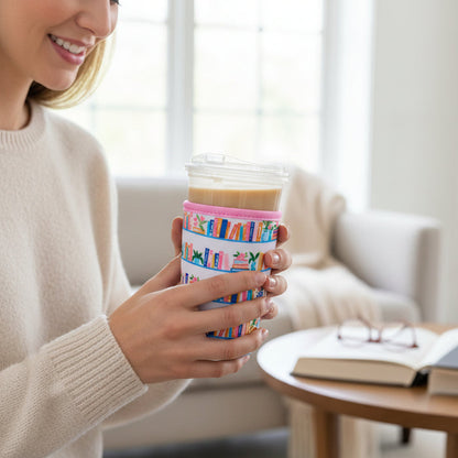 A woman in a cream sweater holds a cup wrapped with The Darling Effect's Large-Book Shelfie Sip Savvy Drink Sleeve. An open book and eyeglasses lie on the wooden table before her, set against a bright living room backdrop.
