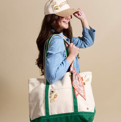 A woman smiles as she adjusts her baseball cap, accessorized with Latte Love The Pin Pack by The Darling Effect. She holds a cream canvas tote with green trim, adorned with collectible accessories and a floral scarf.