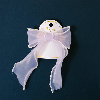 A sheer lilac pearl hair bow with beaded edges is attached to a gold barrette and displayed on a white the-darling-effect card against a dark blue background, adding charming elegance to any look.