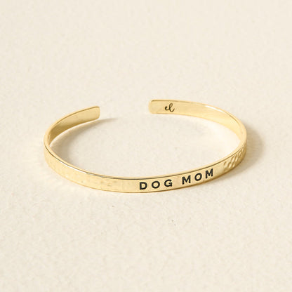 The Darling Effect’s Little Reminder Cuff is a 14k gold plated bracelet engraved with DOG MOM, displayed on a light, textured surface—an ideal gift for any proud pet lover.
