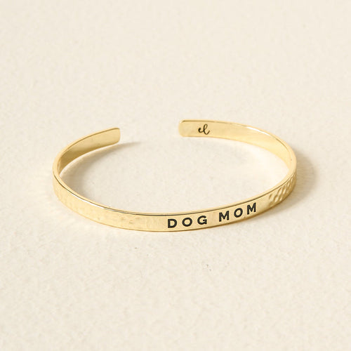 The Darling Effect’s Little Reminder Cuff is a 14k gold plated bracelet engraved with DOG MOM, displayed on a light, textured surface—an ideal gift for any proud pet lover.