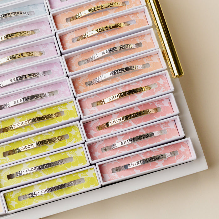 A display tray features The Darling Effect’s Little Reminder Cuff, each engraved with uplifting words like MAMA and SHINE BRIGHT in gold. A 14k gold plated pen rests beside them, making this set a perfect gift.