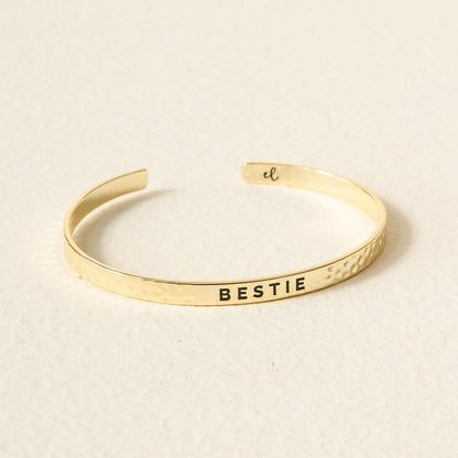 The Darling Effect's Little Reminder Cuff is a 14k gold plated, hammered bracelet engraved with “BESTIE” in black on the front and a small script inside. This gift-ready piece rests on a light, textured surface.