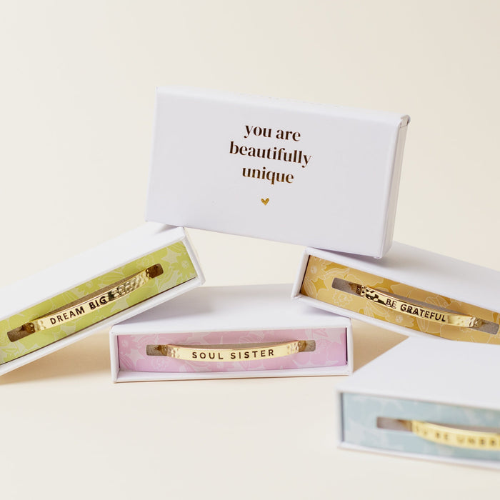 Four Little Reminder Cuffs by The Darling Effect feature “DREAM BIG,” “SOUL SISTER,” and “BE GRATEFUL” messages, plated in 14k gold and packaged in pastel, gift-ready boxes. A white box reads, “you are beautifully unique” in gold.