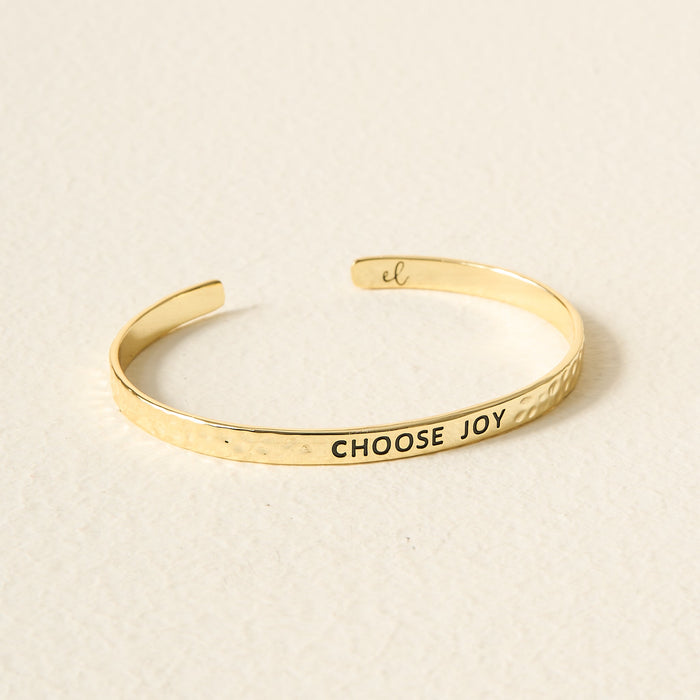 The Darling Effect’s Little Reminder Cuff is a 14k gold plated bracelet with a hammered texture, engraved “CHOOSE JOY” in black capitals—a gift-ready accessory that rests beautifully on any surface.