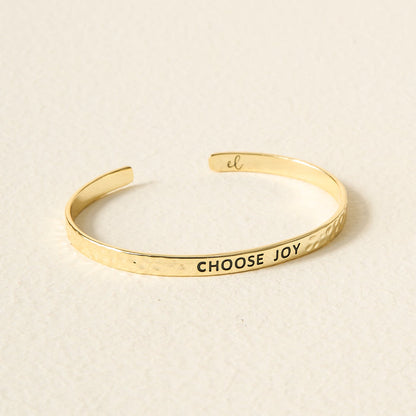 The Darling Effect’s Little Reminder Cuff is a 14k gold plated bracelet with a hammered texture, engraved “CHOOSE JOY” in black capitals—a gift-ready accessory that rests beautifully on any surface.