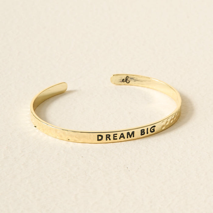 The Darling Effect's Little Reminder Cuff is a 14k gold plated, hammered open bracelet engraved with DREAM BIG outside and a small script inside, beautifully gift-ready on a light textured surface.