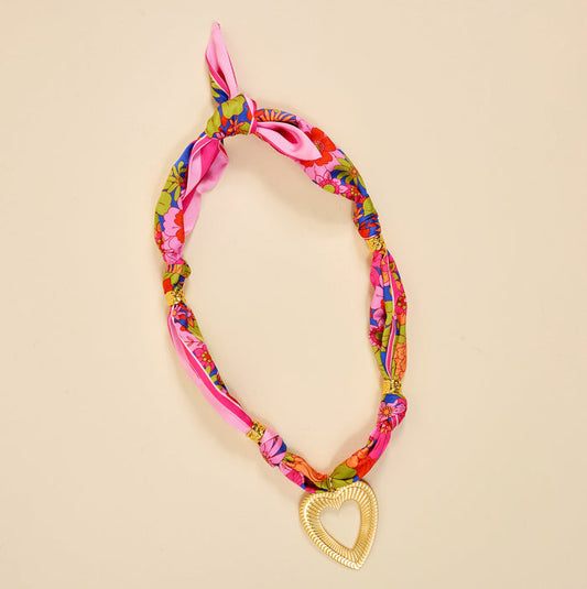 The Darling Effect's Lover's Tempo Make a Statement Scarf Necklace showcases silky pink fabric with colorful florals, finished with a shimmering gold heart pendant—a bold accessory for any look.