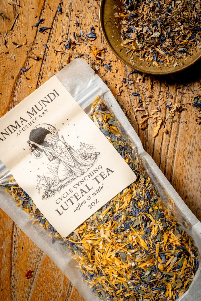 A clear plastic bag of Anima Mundi Apothecary Cycle Swinging Luteal Tea lies on a rustic wooden surface, with a matching bowl of loose tea and scattered dried flowers around it.